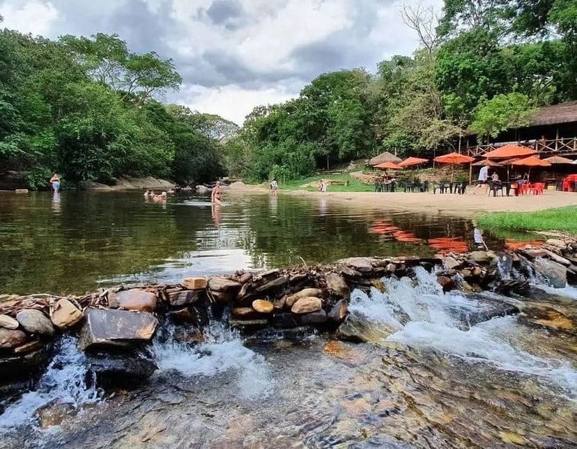Rio de águas rasas com uma pequena queda formada por pedras, rodeado por árvores e com área de lazer com mesas e guarda-sóis ao fundo.