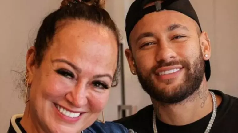 O jogador Neymar ao lado da mãe, ambos usam roupas com tonalidades escuras estão sorrindo.
