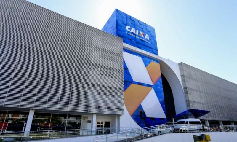 Logotipo da Caixa Econômica Federal em placa sobre fundo azul com céu aberto