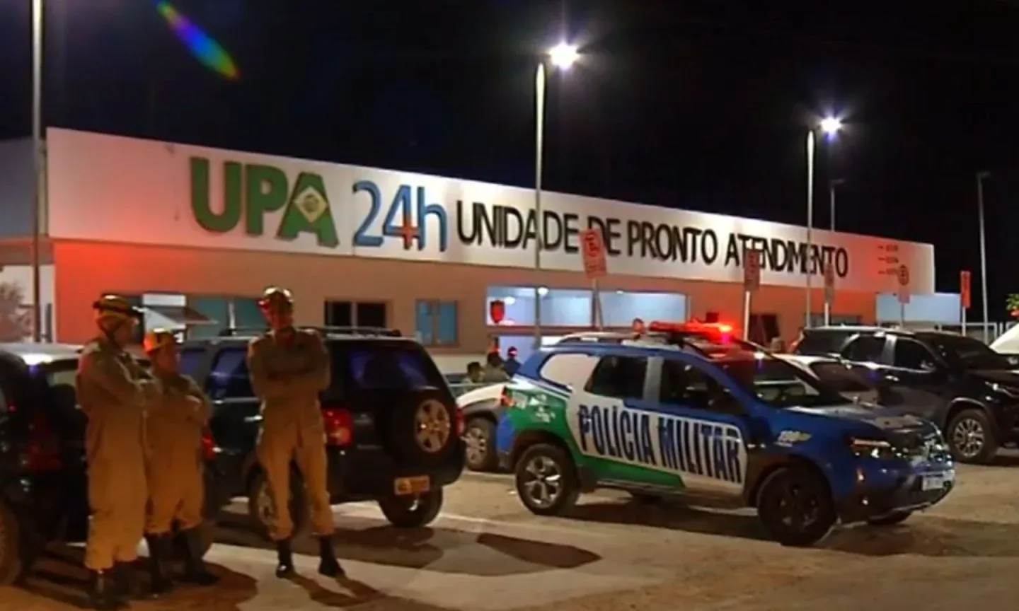 Viatura da Polícia Militar com giroflex ligado e policiais posicionados em frente à UPA 24 horas durante a noite, com o prédio iluminado e movimento de veículos no estacionamento.