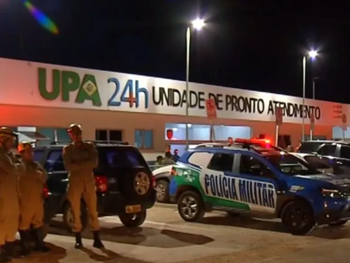 Viatura da Polícia Militar com giroflex ligado e policiais posicionados em frente à UPA 24 horas durante a noite, com o prédio iluminado e movimento de veículos no estacionamento.