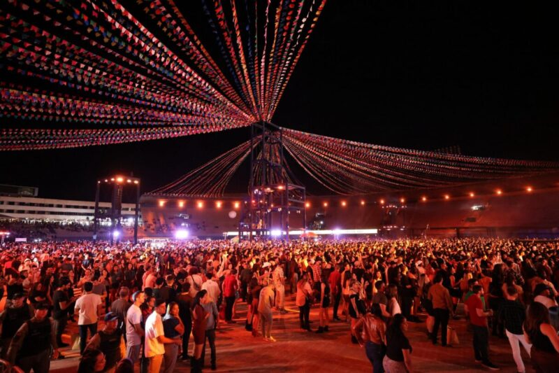 Multidão em um animado arraial noturno, com bandeirolas coloridas e iluminação típica de festa junina.