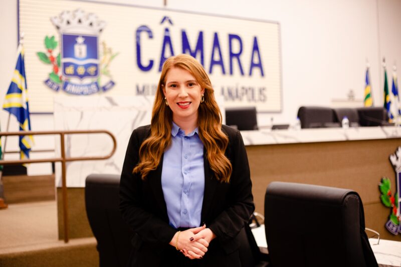 Mulher de cabelos ruivos, usando blazer preto e camisa azul, posa sorrindo em pé no plenário da Câmara Municipal de Anápolis, com o brasão da cidade e a palavra "Câmara" em destaque ao fundo.