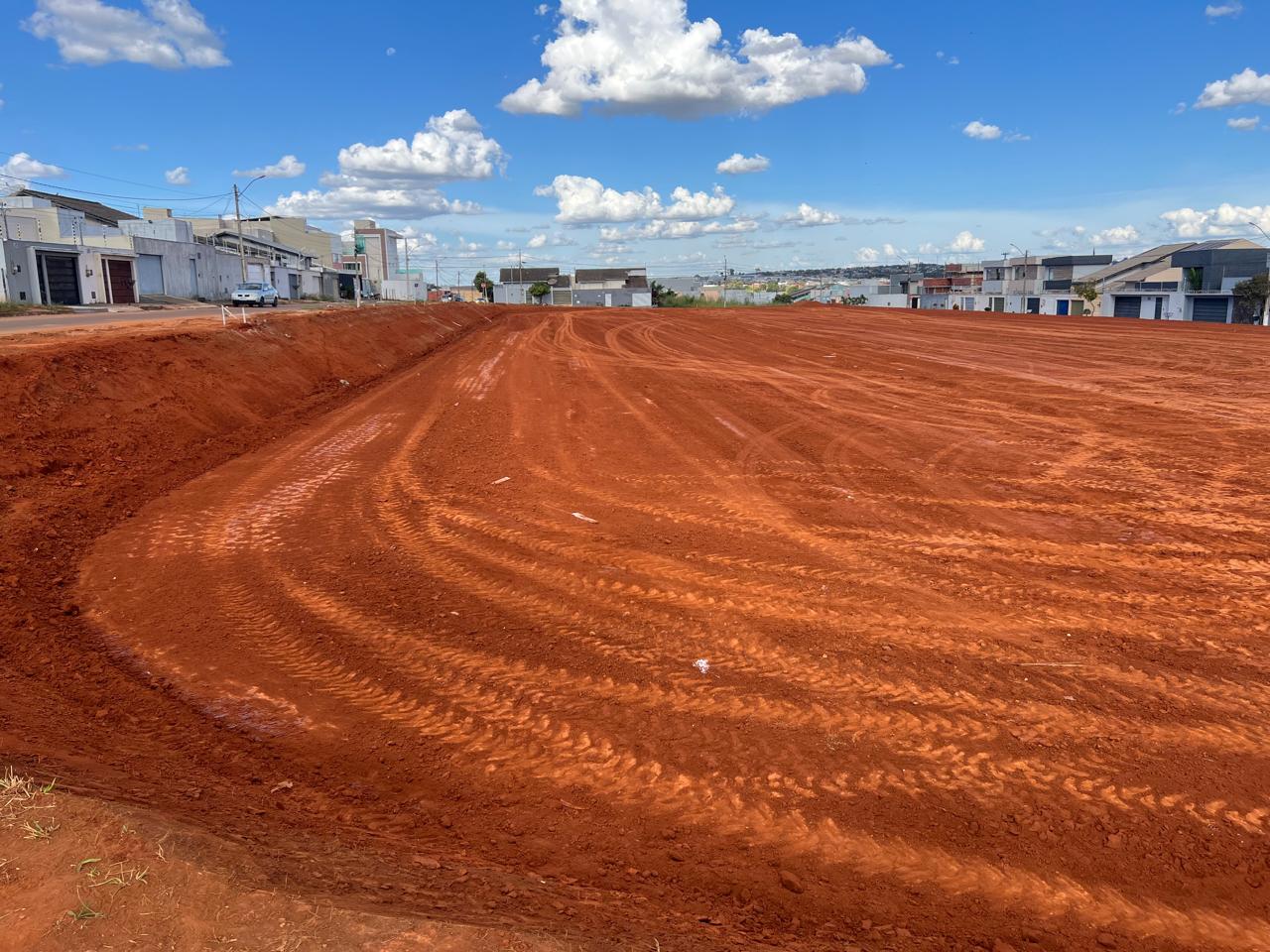 Terreno com solo vermelho em fase de terraplanagem para a construção do novo Colégio Estadual Plínio Jayme, com casas ao fundo e céu azul com nuvens.