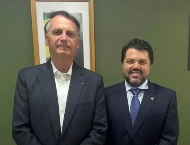 Jair Bolsonaro e Márcio Corrêa posam juntos em um ambiente interno, ambos usando ternos escuros, com um quadro ao fundo.
