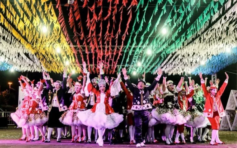 Grupo de homens e mulheres vestidos com trajes típicos de festa junina dançando quadrilha sob bandeirinhas coloridas e vibrantes.