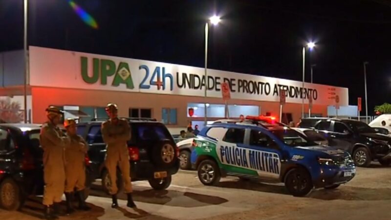 Viatura da Polícia Militar com giroflex ligado e policiais posicionados em frente à UPA 24 horas durante a noite, com o prédio iluminado e movimento de veículos no estacionamento.