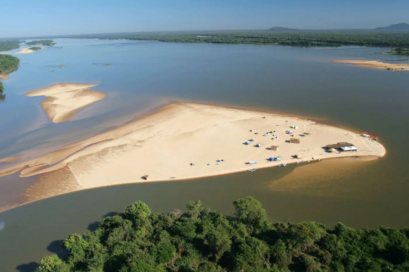 Vista aérea de uma ilha de areia no Rio Araguaia com poucas barracas e mata ao redor.