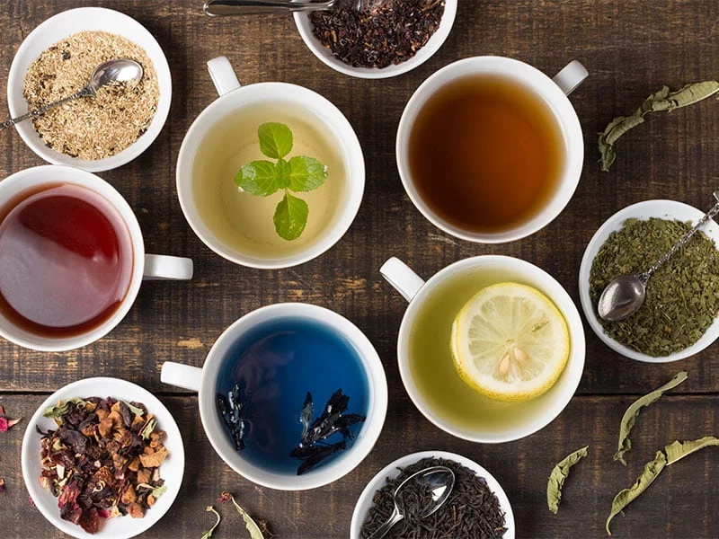Diversas xícaras com tipos variados de chá, incluindo azul, verde, vermelho e amarelo, cercadas por ervas e ingredientes naturais.
