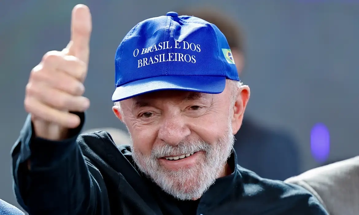 Lula usando boné azul com a frase "O Brasil é dos brasileiros", sorrindo e fazendo sinal de positivo com o polegar.