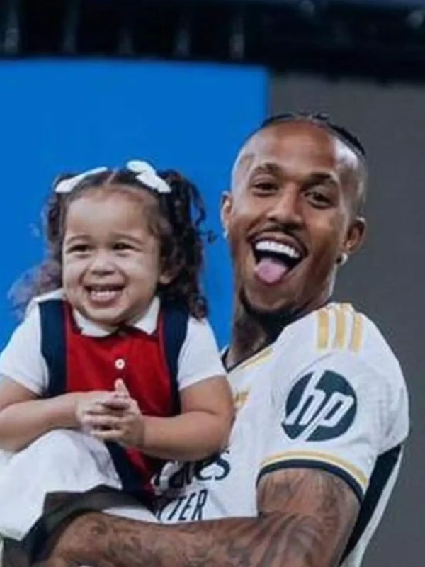 Jogador Éder Militão sorrindo e segurando sua filha Cecília no colo.