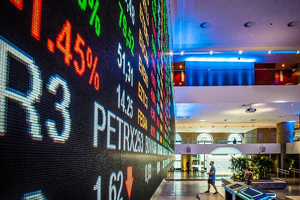 Painel digital da bolsa de valores com cotações de ações em destaque, exibindo números coloridos e índices financeiros em um prédio moderno.