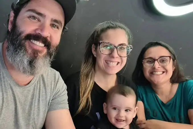 Trisal com bebê posando para foto. Todos estão sorrindo em um ambiente fechado, com clima de alegria e acolhimento familiar.