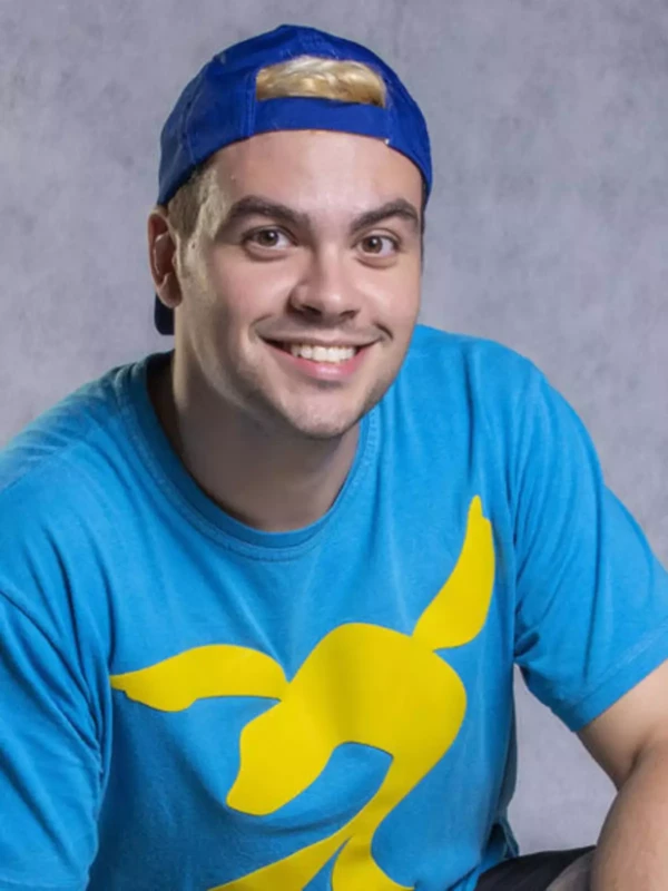 Youtuber Luccas Neto sorri para a câmera vestindo camiseta azul com estampa amarela, cenário lúdico ao fundo