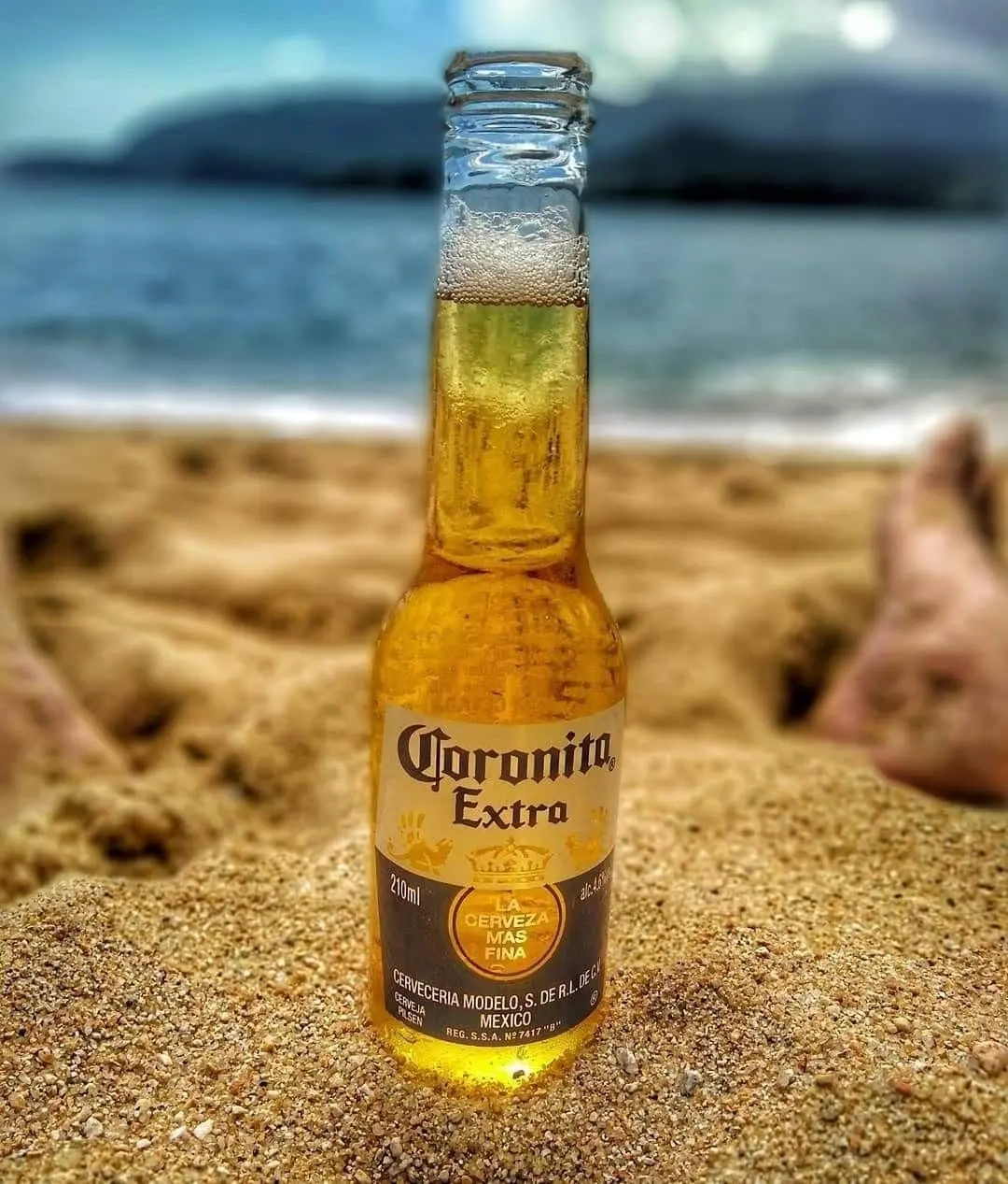 Garrafa de cerveja Coronita Extra na areia da praia, com o mar ao fundo e pés relaxados ao redor.