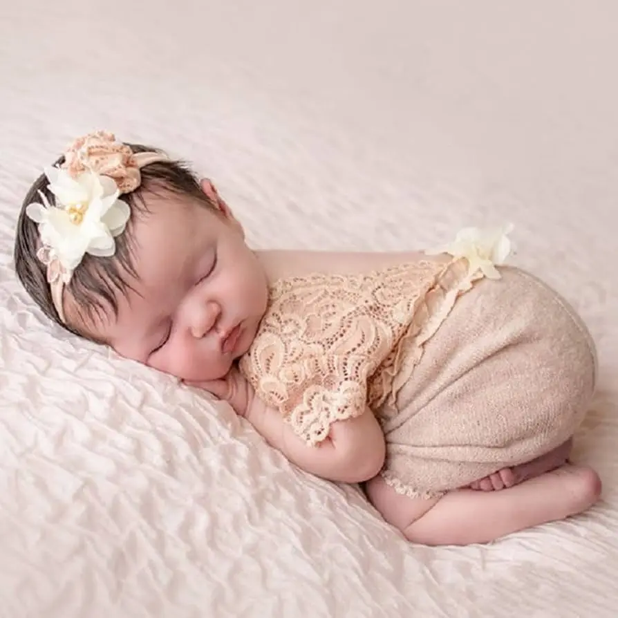 Bebê recém-nascida dormindo de lado com roupa rendada e faixa floral na cabeça.