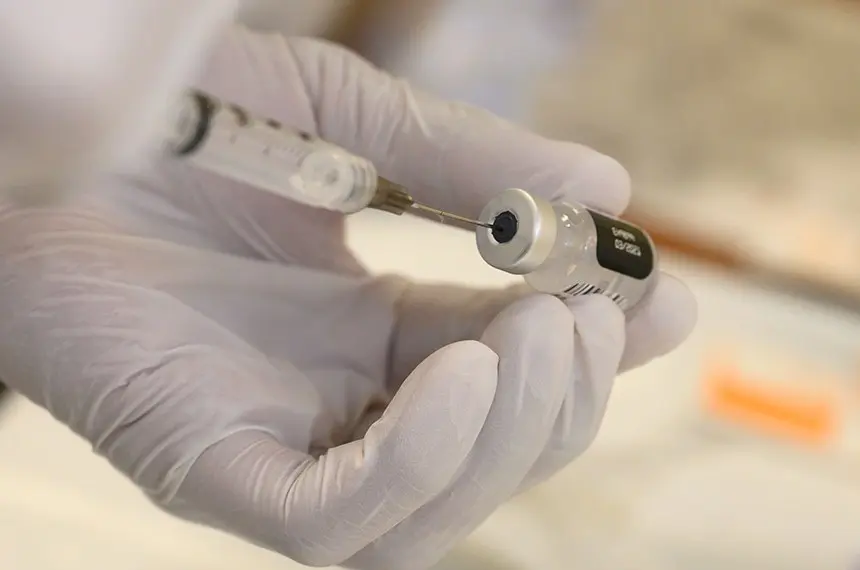 Profissional de saúde com luvas brancas manuseia seringa e frasco de vacina durante preparo da dose.
