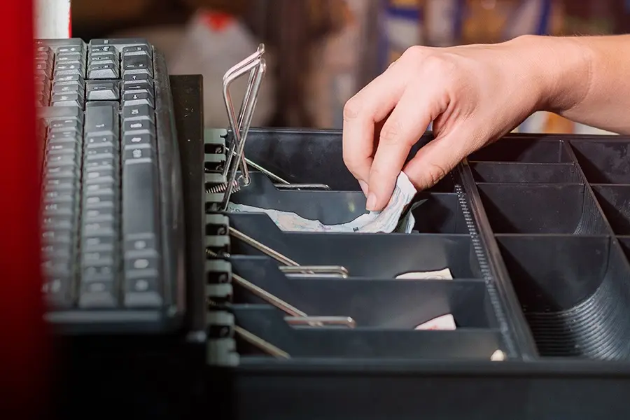Mão retirando cédulas de dinheiro de uma gaveta registradora em caixa de supermercado ao lado de um teclado.