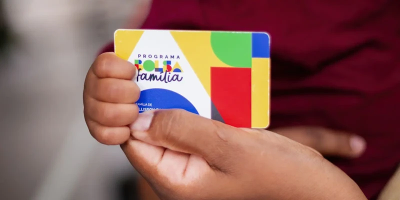 Mulher segurando o cartão do Bolsa Família, com fundo desfocado, representando beneficiária do programa.