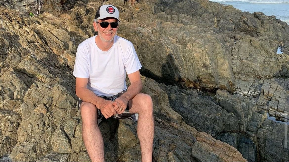 Professor Marco Antônio sentado em rochas, sorrindo, usando camiseta branca e boné