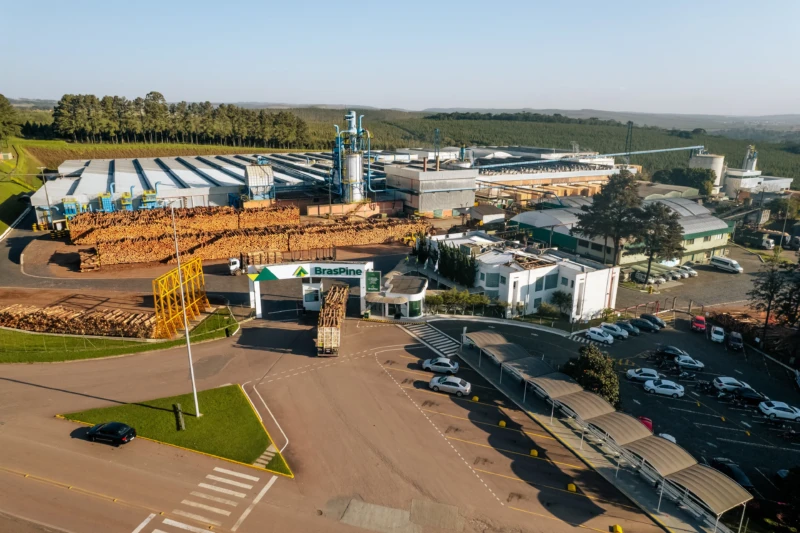 Vista aérea de uma indústria madeireira com grande estrutura, caminhões, pilhas de toras e galpões cercados por vegetação.