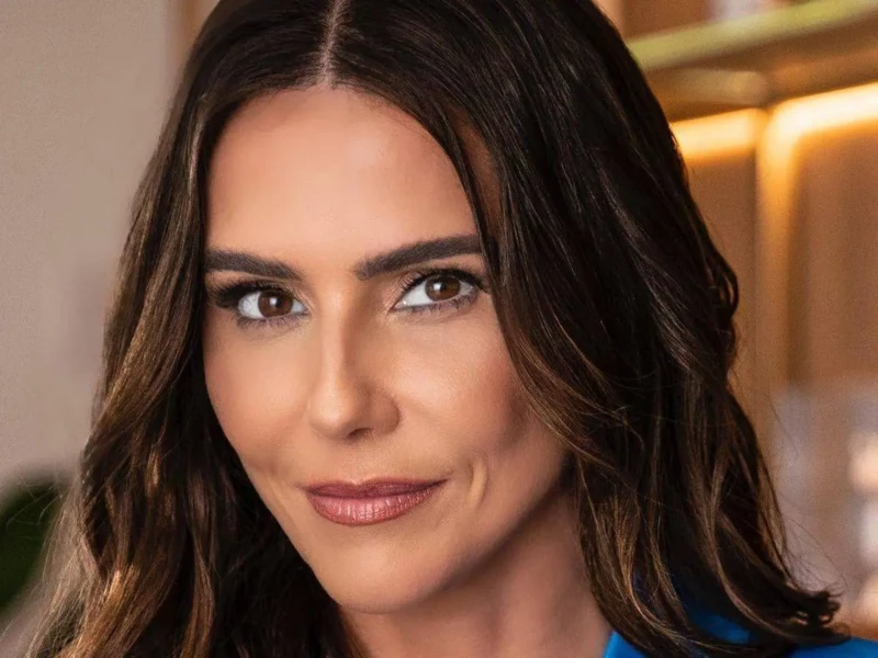 Deborah Secco de frente, com expressão neutra, usando blusa de alça laranja e cabelo solto levemente ondulado, em frente a uma parede azul-claro.