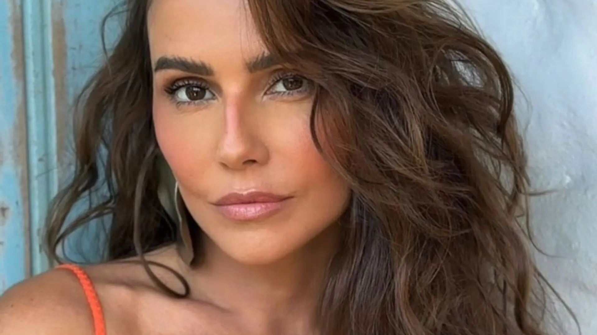 Deborah Secco sorrindo levemente, com cabelos lisos e escuros, vestindo blusa azul, em ambiente interno bem iluminado.