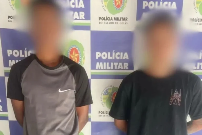 Dois jovens com os rostos desfocados posam diante de um painel da Polícia Militar do Estado de Goiás.