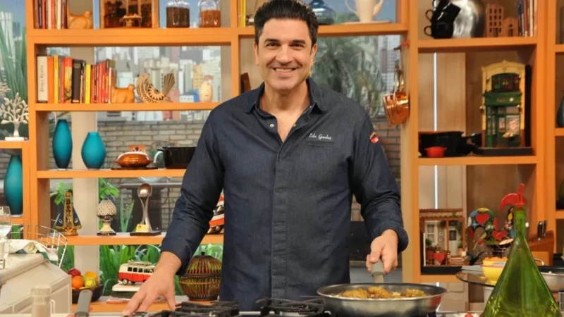 O apresentador Edu Guedes sorrindo enquanto prepara uma refeição em um estúdio de culinária, usando camisa jeans escura.
