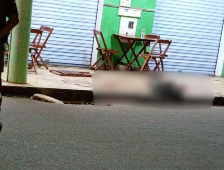 Imagem desfocada mostra corpo caído na calçada em frente a uma sorveteria, com mesas e cadeiras vazias.