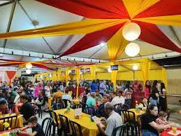 Pessoas sentadas e em pé participam da festa da Divino Pai Eterno sob uma tenda com decoração vermelha e amarela.