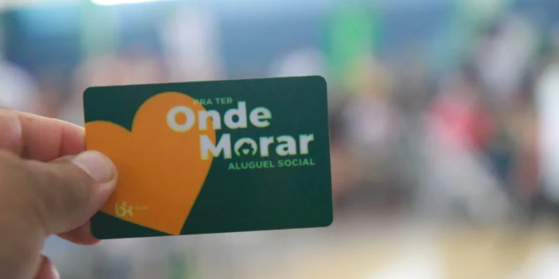 Pessoa segura cartão do Aluguel Social com logo do programa e coração laranja