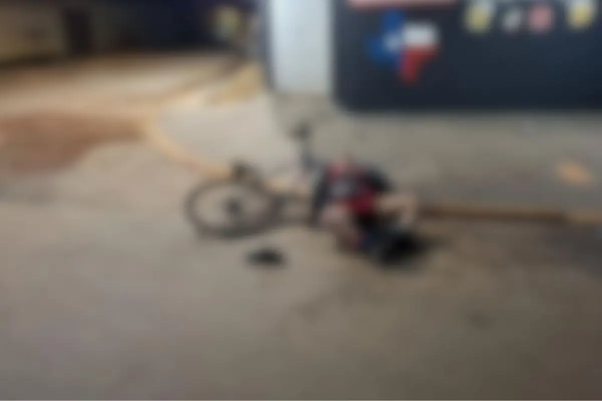 Corpo de um homem caído ao lado de uma bicicleta em uma rua escura e deserta, imagem desfocada.