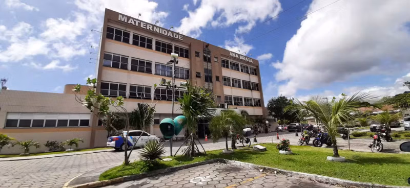Vista externa da Maternidade Balbina Mestrinho, com céu azul e nuvens, árvores e pessoas circulando na área externa.