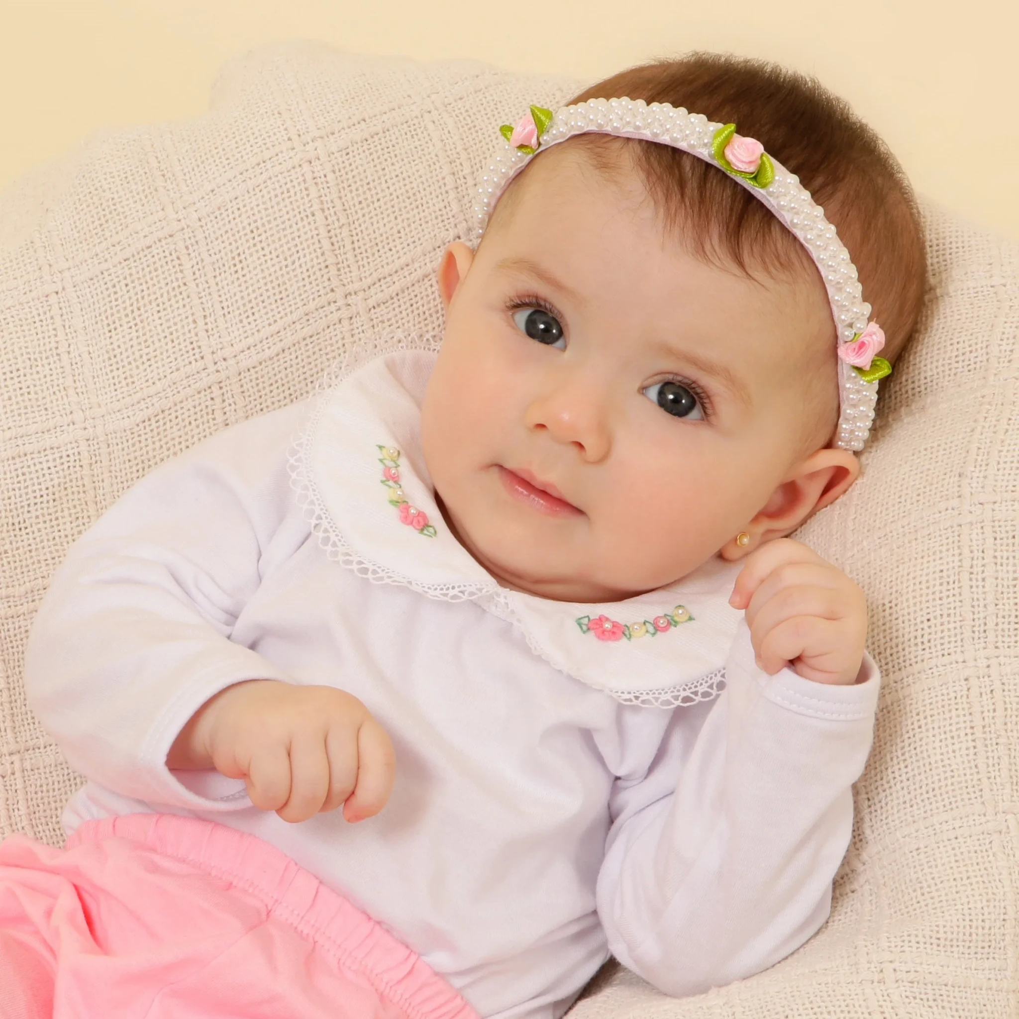 Bebê com olhos grandes e expressivos, vestida de branco e rosa, com tiara de pérolas e flores.
