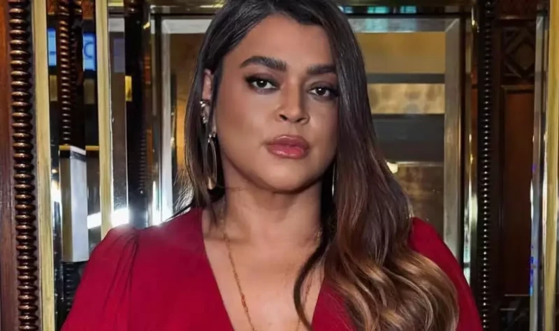 Cantora Preta Gil sorri usando vestido preto com transparência nos ombros; ela está com cabelo solto e maquiagem leve, em fundo neutro.