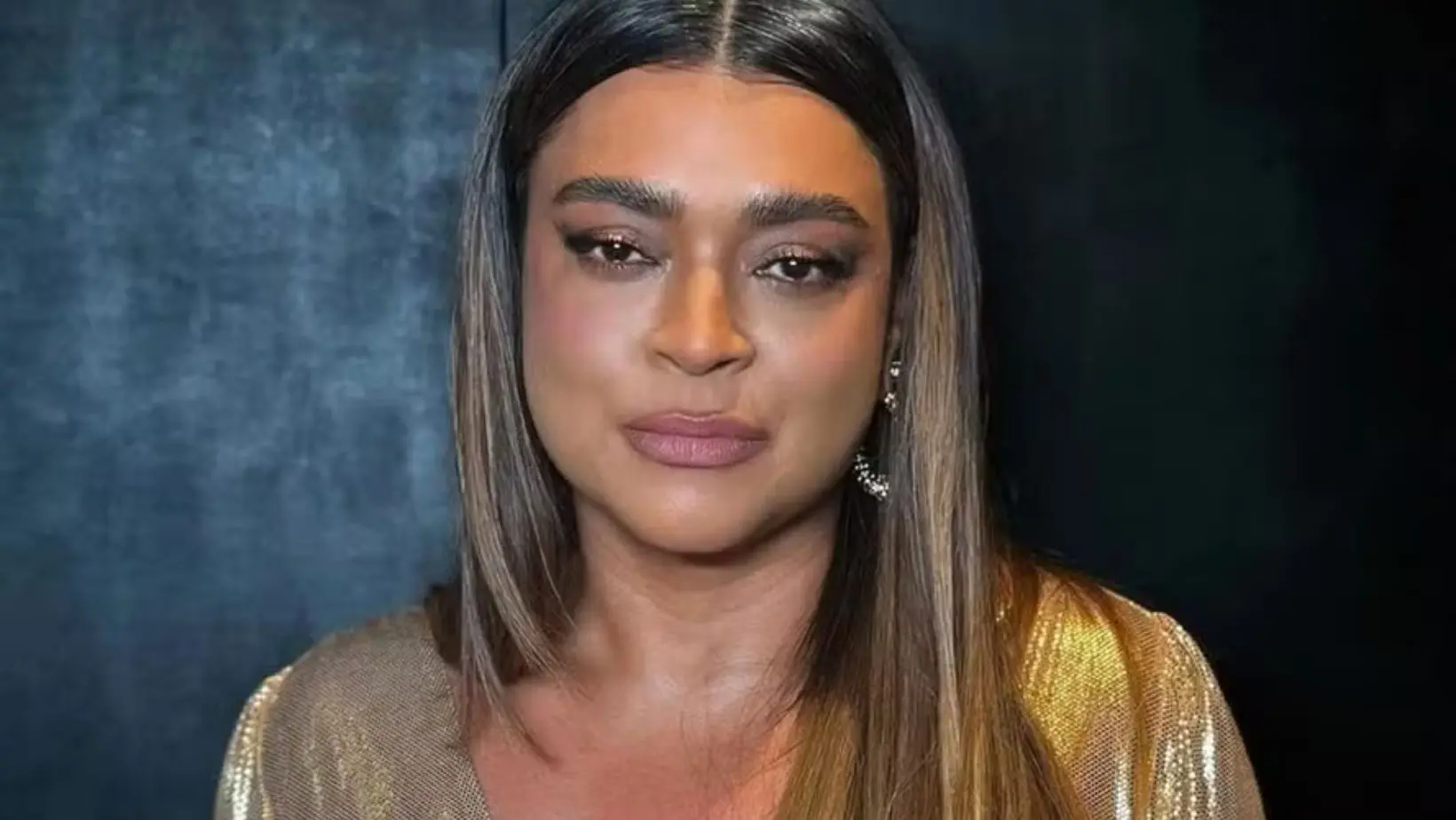 Preta Gil em apresentação musical, usando figurino dourado e sorrindo para o público no palco, com iluminação artística ao fundo.