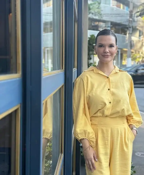 Empresária Natália Thomazella em pé, usando conjunto amarelo, próxima a uma parede de vidro