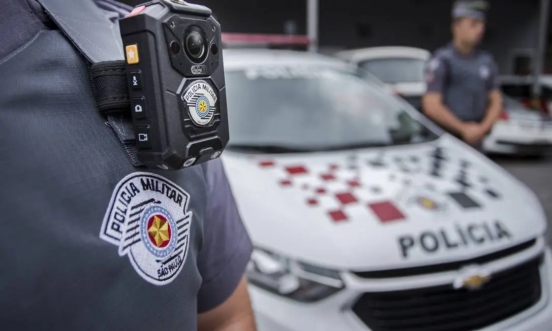 Policial militar de São Paulo usa câmera corporal ao lado de uma viatura da corporação.