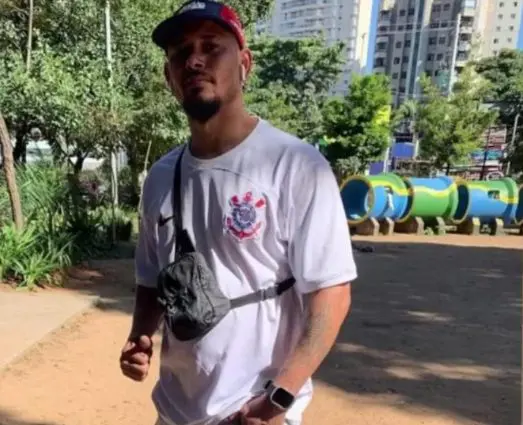 Homem usando camiseta branca e boné em uma área pública ao ar livre.