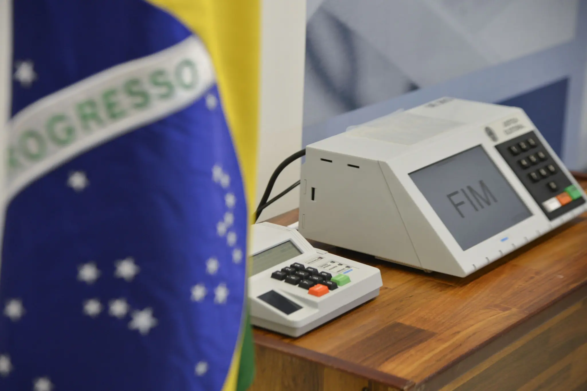 Urna eletrônica utilizada nas eleições brasileiras sobre mesa, com bandeira do Brasil ao fundo. Foto: Nelson Jr./Asics/TSE.