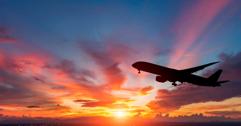 Avião comercial sobrevoa o céu durante o pôr do sol, em imagem ilustrativa sobre viagens aéreas e milhas.