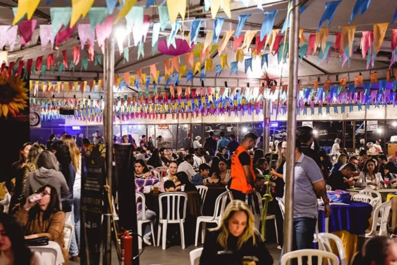 Público participa de festa com barraquinhas, mesas e decoração com bandeirinhas coloridas.