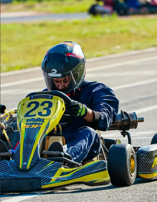 Piloto de capacete e macacão azul conduz kart amarelo e azul, identificado pelo número 23, durante corrida em pista.