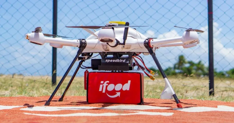 Drone de entregas do iFood com compartimento vermelho para transporte de pedidos, posicionado em uma área de pouso.