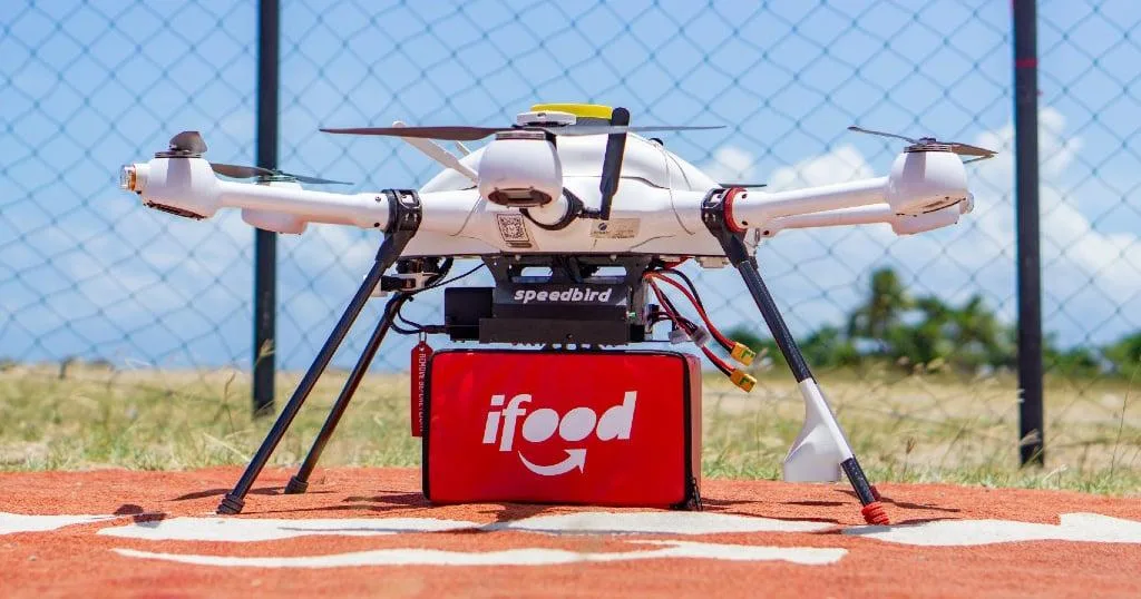 Drone de entregas do iFood com compartimento vermelho para transporte de pedidos, posicionado em uma área de pouso.