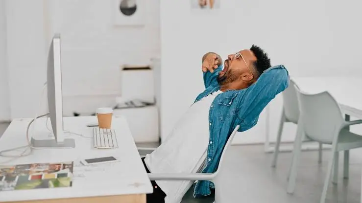 Homem boceja enquanto trabalha em frente ao computador, em imagem ilustrativa sobre sonolência durante o dia.