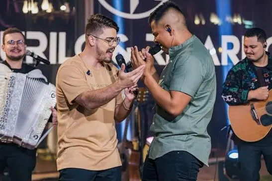 Dois cantores se apresentam no palco com microfones, acompanhados por músicos com sanfona e violão.