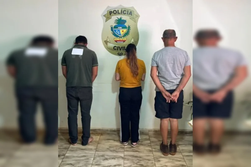 Três pessoas aparecem de costas diante do símbolo da Polícia Civil de Goiás, em uma unidade policial.