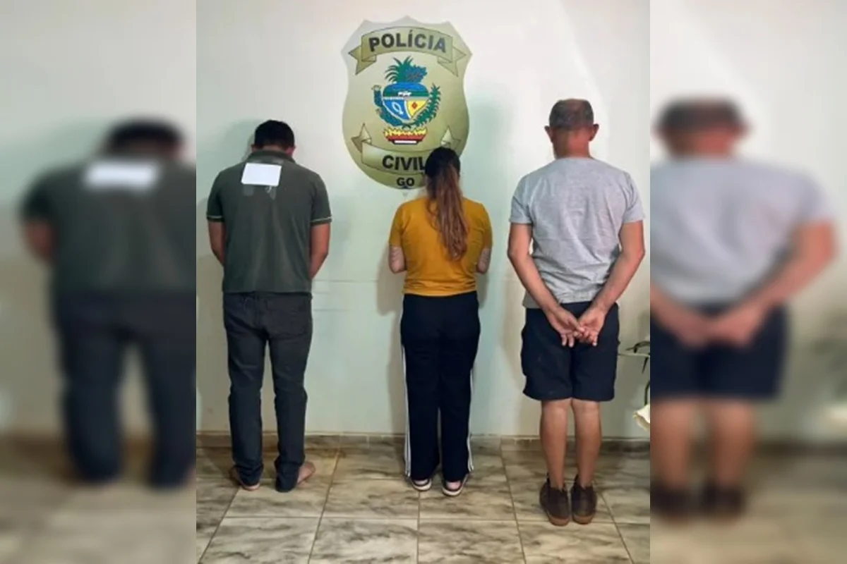 Três pessoas aparecem de costas diante do símbolo da Polícia Civil de Goiás, em uma unidade policial.